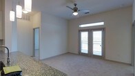 This single-floor residence features an open-concept living area finished with neutral-toned carpet and light-colored walls that create a bright, clean atmosphere. The main living space is anchored by a ceiling fan with an integrated light fixture, providing both air circulation and ambient lighting. A set of French doors serves as a focal point, offering direct access to a private balcony and allowing natural light to fill the room through both the doors and a transom window above. The balcony beyond the doors suggests outdoor views and additional ventilation options. A granite countertop is partially visible at the edge of the frame, indicating an adjacent kitchen or breakfast bar area that transitions seamlessly into the living space. A doorway visible toward the rear of the unit leads to additional rooms, suggesting a practical layout that separates living and private areas. The overall design is simple and functional, with neutral finishes throughout that provide a versatile backdrop suitable for a variety of interior styles. The space presents a comfortable, well-lit environment with a balance of indoor living and outdoor connectivity.