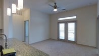 This single-floor residence features an open-concept living area finished with neutral-toned carpet and light-colored walls that create a bright, clean atmosphere. The main living space is anchored by a ceiling fan with an integrated light fixture, providing both air circulation and ambient lighting. A set of French doors serves as a focal point, offering direct access to a private balcony and allowing natural light to fill the room through both the doors and a transom window above. The balcony beyond the doors suggests outdoor views and additional ventilation options. A granite countertop is partially visible at the edge of the frame, indicating an adjacent kitchen or breakfast bar area that transitions seamlessly into the living space. A doorway visible toward the rear of the unit leads to additional rooms, suggesting a practical layout that separates living and private areas. The overall design is simple and functional, with neutral finishes throughout that provide a versatile backdrop suitable for a variety of interior styles. The space presents a comfortable, well-lit environment with a balance of indoor living and outdoor connectivity.