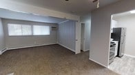 This single-floor residence offers a thoughtfully organized layout with a variety of functional spaces. The galley-style kitchen stands out as a highlight, featuring crisp white shaker cabinetry, marble-style countertops, a double stainless steel sink, and a full suite of appliances including a gas range and refrigerator. Light wood-look flooring runs throughout the kitchen, lending a clean, contemporary feel.

The living areas are finished with warm, neutral-toned carpet and painted in soft gray and beige tones, creating a calm, cohesive atmosphere throughout. Exposed ceiling beams and a ceiling fan add character to the main living space, while vertical shiplap-style paneling on a central dividing wall provides a subtle architectural detail. Baseboard heating and wall-mounted air conditioning units are present throughout the carpeted rooms, ensuring year-round comfort.

The bedrooms are generously proportioned, each with carpet flooring, windows allowing natural light, and ample wall space. One bedroom features a dedicated vanity area with a granite-style countertop and mirror, situated adjacent to a walk-in closet with built-in shelving. The full bathroom is finished with tile surround, a soaking tub and shower combination, a white vanity, and wood-look flooring, offering a clean and practical design.