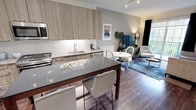 This contemporary single-floor residence showcases a thoughtfully designed open-concept layout that seamlessly connects the kitchen, dining, and living areas. The kitchen is a standout feature, outfitted with light wood-grain cabinetry extending to the ceiling, stainless steel appliances including a gas range, microwave, and French door refrigerator, white tile backsplash, and a large central island with a sleek dark quartz countertop and bar seating. Rich dark hardwood flooring runs throughout the main living area, adding warmth and contrast to the neutral palette. The living room is bright and inviting, anchored by sliding glass doors that open to a private balcony with lush outdoor views, allowing generous natural light to fill the space. The bedroom offers a serene retreat with plush carpet, soft gray accent walls, and large windows that frame garden views. A striking channeled velvet headboard serves as a focal point, while an adjoining walk-in closet with built-in shelving provides ample storage. The bathroom is finished in a clean, minimalist style with a warm wood vanity, white quartz countertop, framed mirror, and bright lighting — creating a crisp, spa-like atmosphere. Throughout the residence, a cohesive modern aesthetic is maintained with neutral tones, quality finishes, and functional design.