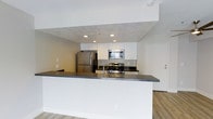 This single-floor 1-bedroom, 1-bathroom residence is designed with a clean, contemporary aesthetic that maximizes both functionality and comfort. The open-concept kitchen serves as the heart of the home, featuring sleek white cabinetry, stainless steel appliances including a gas range, built-in microwave, and a full-size refrigerator. A broad kitchen peninsula with a dark countertop provides ample prep space and doubles as a casual dining or entertaining surface. Recessed lighting illuminates the kitchen area, highlighting the crisp white finishes and modern hardware. The flooring throughout features light-toned wood-look planks that add warmth while maintaining a contemporary feel. Smooth white walls and neutral tones create a bright, airy atmosphere across the living areas. The layout flows efficiently from the kitchen into the living space, making the most of every square foot. The entry door is positioned conveniently off the main living area, contributing to a logical and practical floor plan. Overall, this residence presents a polished, move-in-ready interior suited for those seeking a modern, low-maintenance living environment.