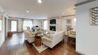 This thoughtfully designed single-floor space offers a warm and inviting atmosphere anchored by a striking floor-to-ceiling stone fireplace, which serves as the centerpiece of the main lounge area. Rich hardwood flooring flows seamlessly throughout, complementing the neutral, light-toned walls and creating a cohesive, elegant aesthetic.

The main lounge is generously proportioned, furnished with comfortable cream sofas and accent chairs arranged around a central coffee table and area rug, encouraging relaxed conversation and gathering. Recessed lighting illuminates the space evenly, while multiple windows dressed with classic blinds and drape panels allow ample natural light to filter in, enhancing the open and airy feel.

Adjacent to the lounge, a bar-height dining counter with dark wood stools provides a casual seating option, seamlessly connecting to a glimpse of a well-appointed kitchen beyond, featuring dark cabinetry, pendant lighting, and modern appliances.

A separate dining nook, defined by a substantial round wood table and leather chairs, offers a more intimate setting and benefits from direct access to the outdoors through glass-paneled doors. Decorative wall art and mirrors add character and warmth throughout. The space is well-suited for community gathering, with a welcoming entry area featuring a cozy sofa and framed artwork that sets a refined, comfortable tone from the moment of arrival.