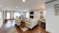 This thoughtfully designed single-floor space offers a warm and inviting atmosphere anchored by a striking floor-to-ceiling stone fireplace, which serves as the centerpiece of the main lounge area. Rich hardwood flooring flows seamlessly throughout, complementing the neutral, light-toned walls and creating a cohesive, elegant aesthetic.

The main lounge is generously proportioned, furnished with comfortable cream sofas and accent chairs arranged around a central coffee table and area rug, encouraging relaxed conversation and gathering. Recessed lighting illuminates the space evenly, while multiple windows dressed with classic blinds and drape panels allow ample natural light to filter in, enhancing the open and airy feel.

Adjacent to the lounge, a bar-height dining counter with dark wood stools provides a casual seating option, seamlessly connecting to a glimpse of a well-appointed kitchen beyond, featuring dark cabinetry, pendant lighting, and modern appliances.

A separate dining nook, defined by a substantial round wood table and leather chairs, offers a more intimate setting and benefits from direct access to the outdoors through glass-paneled doors. Decorative wall art and mirrors add character and warmth throughout. The space is well-suited for community gathering, with a welcoming entry area featuring a cozy sofa and framed artwork that sets a refined, comfortable tone from the moment of arrival.