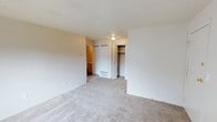 This single-floor, one-bedroom, one-bathroom unit is designed for comfortable, efficient living. The bedroom is a well-proportioned space featuring soft, neutral-toned wall-to-wall carpeting that adds warmth underfoot. A textured ceiling with a centrally mounted flush light fixture provides general illumination throughout the room. Storage is well-addressed with two closets: one standard closet and one open-style closet with visible shelving and a hanging rod, offering ample organization options. The neutral, light-toned walls create a clean and versatile backdrop suitable for a variety of décor styles. A floor vent indicates a forced-air climate control system for year-round comfort. Visible from the bedroom is a connected hallway leading to the bathroom, where warm-toned cabinetry can be glimpsed, suggesting functional vanity storage. The overall palette of the unit is consistent and cohesive, with beige and off-white tones throughout, lending the space a calm, neutral ambiance. This unit is an efficient and practical layout suited for single occupants or couples seeking straightforward, manageable living quarters.