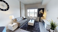 Located at 229 Andover Park E in Tukwila, Washington, this modern single-floor unit showcases a clean, contemporary design throughout. The living area is an inviting, open space highlighted by soaring textured ceilings, light hardwood-style flooring, and large windows that flood the room with natural light while offering views of the surrounding cityscape and greenery. The living space is anchored by a neutral color palette, with crisp white walls complemented by bold concrete-accented columns and ceiling detailing that lend an industrial-chic character to the interior. The kitchen is thoughtfully designed with a striking two-tone cabinetry scheme — deep navy lower cabinets contrasted by bright white upper cabinets — paired with sleek white quartz countertops and a full suite of stainless steel appliances, including a refrigerator, electric range, over-the-range microwave, and dishwasher. Track lighting illuminates the workspace efficiently. The bathroom continues the refined aesthetic with navy cabinetry, a white countertop vanity, an LED-backlit mirror, and wood-look tile flooring, creating a clean and polished feel. Light hardwood-style flooring flows consistently throughout the unit, tying each space together in a cohesive and modern design.
