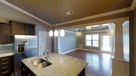 This single-floor residence showcases a thoughtfully designed open-concept layout that seamlessly connects the kitchen and living areas. The kitchen is anchored by a substantial granite-topped island with a double undermount sink and a modern gooseneck faucet, complemented by stainless steel appliances and dark cabinetry. A trio of pendant lights hang above the island, providing focused task lighting with a stylish touch. The kitchen transitions into the living and dining area via decorative white columns and a half-wall, adding architectural character while maintaining an open, airy feel. The living area features rich hardwood flooring and is distinguished by a coffered tray ceiling with recessed lighting, lending a sense of elegance and dimension to the space. Neutral gray walls throughout provide a versatile backdrop that pairs well with the warm wood tones and white trim. A large window and glass door in the living area invite abundant natural light and offer views of the outdoors. Crown molding and crisp white baseboards add refined finishing details throughout, giving the space a polished, cohesive aesthetic overall.