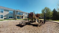 This single-story residential community offers a welcoming outdoor environment centered around a dedicated playground area. The play space features a multi-level climbing structure equipped with a dual-lane slide, multiple access points, and overhead bars, all set atop a wood chip safety surface that spans the activity zone. A picnic table with attached bench seating is positioned nearby, providing a convenient spot for supervision or casual outdoor gatherings.

The surrounding buildings display a traditional architectural style, characterized by neutral siding in soft yellow tones, pitched rooflines, and covered entryways. Multiple windows are visible across the façades, allowing natural light into the units and offering views of the outdoor common area. Mature deciduous trees with seasonal foliage in warm autumn tones are scattered throughout the grounds, adding color and shade to the landscape.

The community layout appears designed with resident interaction in mind, placing the play area in a central, accessible location visible from the surrounding buildings. A paved pathway connects the play space to the broader community, enhancing walkability. Under clear blue skies, the outdoor amenities create an inviting and family-friendly atmosphere suitable for everyday recreation.