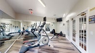 Located at 8700 Tierra Vista Cir in Kissimmee, Florida, this fitness center offers a bright, well-appointed space designed to support a complete exercise regimen. The room features light-toned hardwood-style flooring that lends a clean, modern aesthetic throughout. Large, multi-pane French doors and windows line the walls, flooding the interior with natural light and providing views of the surrounding outdoor areas, creating an open and inviting atmosphere.

The space is equipped with a solid selection of cardio equipment, including a treadmill, elliptical trainer, and stair stepper, each positioned to allow comfortable use. A dedicated free weights area features a rack of dumbbells in varying sizes alongside an adjustable workout bench, catering to strength training needs. Full-length wall mirrors span a significant portion of the room, visually expanding the space and allowing users to monitor their form during workouts.

A wall-mounted flat-screen television provides entertainment during sessions, while motivational signage adds an energetic touch to the décor. A stability ball and additional accessories are available for flexibility and supplemental training. Fluorescent lighting ensures the room remains well-lit throughout the day. The overall layout is efficiently organized, making the most of the available footprint while maintaining clear pathways between equipment.