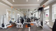 This fitness center is designed to accommodate a full range of workout needs within a well-appointed, single-level space. The room features a tray ceiling with crown molding, recessed lighting, and ceiling fans that contribute to a comfortable exercise environment. Large windows fitted with horizontal blinds line the walls, allowing generous natural light to filter in while offering views of an adjacent outdoor pool area. A glass door provides direct access to the exterior.

The flooring consists of a patterned carpet tile in grey and orange, providing both durability and visual interest throughout the space. Full-length mirrors are mounted along one wall, visually expanding the room and allowing users to monitor their form during exercise.

The cardio section is equipped with treadmills, elliptical trainers, and upright bikes. The strength training area includes multiple Quantum cable machines, a power tower for bodyweight exercises, free-weight benches, a dumbbell rack, and additional cable stations positioned along the perimeter. Wall-mounted flat-screen televisions are placed at strategic locations for viewing during workouts. The overall layout is organized to allow easy circulation and simultaneous use of multiple equipment stations.