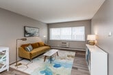 This single-floor unit offers a practical and well-organized layout across several distinct spaces. The open-concept living and dining area forms the heart of the unit, finished with warm wood-look laminate flooring that extends throughout the main living spaces. A large window allows natural light to fill the room, complementing the neutral gray walls and textured ceiling. The living area provides ample room for seating and a dining setup, making it a versatile and welcoming space.

The kitchen is positioned adjacent to the living area and features warm oak-toned cabinetry, dark laminate countertops, a dishwasher, and a full-size refrigerator. The continued wood-look flooring ties the kitchen visually to the rest of the unit.

The primary bedroom is carpeted and includes a ceiling fan, a wall-unit air conditioner, a window with exterior views, and an open closet with shelving for storage. A second bedroom, also carpeted and equipped with a ceiling fan and window, accommodates two beds, making it suitable for shared occupancy.

The bathroom features a white vanity with under-cabinet storage, a large mirror, chrome fixtures, and a shower-tub combination. Overall, the unit presents a clean, functional aesthetic with consistent finishes throughout.