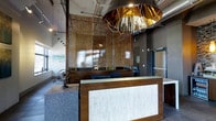 This single-floor space makes an immediate impression with its thoughtfully curated, industrial-chic aesthetic. The reception area serves as the focal point, anchored by a striking wooden desk with an illuminated textured front panel that lends a warm, welcoming glow. Overhead, a dramatic oversized pendant light with a copper-toned metallic interior commands attention, acting as a bold artistic statement that sets the tone for the entire space.

The design layers materials with intention — warm wood tones contrast with cool concrete-look surfaces, while an ornate metallic mesh screen partition adds depth and visual intrigue, subtly dividing the front area from the workspace beyond. Exposed ductwork and industrial ceiling details reinforce the space's contemporary character, while track lighting provides practical, well-distributed illumination throughout.

Decorative accents, including a sculptural armillary sphere, contribute to the space's refined personality, suggesting a professional environment with a creative edge. Natural light filters in through windows, complementing the warm artificial lighting. Overall, the space balances functionality with distinctive design, creating an environment that is both highly professional and visually memorable.