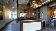 This single-floor space makes an immediate impression with its thoughtfully curated, industrial-chic aesthetic. The reception area serves as the focal point, anchored by a striking wooden desk with an illuminated textured front panel that lends a warm, welcoming glow. Overhead, a dramatic oversized pendant light with a copper-toned metallic interior commands attention, acting as a bold artistic statement that sets the tone for the entire space.

The design layers materials with intention — warm wood tones contrast with cool concrete-look surfaces, while an ornate metallic mesh screen partition adds depth and visual intrigue, subtly dividing the front area from the workspace beyond. Exposed ductwork and industrial ceiling details reinforce the space's contemporary character, while track lighting provides practical, well-distributed illumination throughout.

Decorative accents, including a sculptural armillary sphere, contribute to the space's refined personality, suggesting a professional environment with a creative edge. Natural light filters in through windows, complementing the warm artificial lighting. Overall, the space balances functionality with distinctive design, creating an environment that is both highly professional and visually memorable.