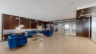 La Citadelle, located at 2125 Rue St Marc in Montreal's vibrant Shaughnessy Village neighbourhood, presents a polished and functional ground-floor lobby that serves as the welcoming gateway to the building. The space is thoughtfully organized into distinct zones, encompassing a main lounge and seating area, a front entrance vestibule with street visibility, and a service corridor leading to the elevator bank and mailroom.

The main lounge is elegantly appointed with rich dark wood wall paneling, recessed pot lighting, and large-format beige tile flooring that extends throughout. Comfortable royal blue upholstered sofas and armchairs are arranged around a gold-accented coffee table, creating an inviting and refined atmosphere for residents and guests. A frosted glass partition subtly divides the seating area from the circulation space, adding a layer of sophistication.

The entrance vestibule features full-height glazing and glass doors that flood the area with natural light and provide an open view to the streetscape. A glazed management or reception office is situated adjacent to the entrance, offering convenient on-site support. The elevator lobby houses multiple stainless steel elevator doors framed by dark wood accents and recessed ceiling details, with a nearby reception counter and mailroom completing the building's comprehensive ground-floor amenities.