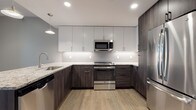 This single-floor residence showcases a thoughtfully designed layout that balances modern finishes with functional living spaces. The kitchen serves as a standout feature, boasting a two-tone cabinet scheme that pairs crisp white upper cabinets with dark wood-grain lower cabinets. Granite countertops, a penny-tile backsplash with under-cabinet lighting, and a full suite of stainless steel appliances—including a range, microwave, dishwasher, and refrigerator—complete the space. Light wood-style flooring runs throughout the kitchen and living areas, contributing to a warm, cohesive aesthetic.

The open living area benefits from natural light through white-trimmed windows and flows seamlessly toward the kitchen. The bedroom features plush wall-to-wall carpeting, dual closets with paneled doors, and corner windows that invite natural light. Neutral taupe tones throughout create a calm, welcoming atmosphere.

The bathroom is finished with large-format tile flooring, a dark wood vanity with a stone-topped countertop, and a glass-enclosed shower. An in-unit laundry area accommodates a stacked washer and dryer, adding everyday convenience to this well-appointed residence.