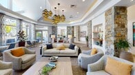 This single-story community clubhouse is defined by a sophisticated blend of natural and contemporary design elements. The entry features large-format tile flooring, a skylight ceiling that floods the space with natural light, floor-to-ceiling glass panels, and prominent stacked-stone columns that establish a warm, resort-inspired aesthetic carried throughout the facility.

The expansive lounge area serves as the centerpiece, showcasing a coffered ceiling with gold-accent cove lighting and striking spiral pendant fixtures. Multiple seating arrangements—including neutral sofas, leather armchairs, and accent chairs—are organized around a linear fireplace and wall-mounted television. Built-in display niches and curated artwork contribute to a polished, gallery-like atmosphere.

Adjacent to the lounge, a well-equipped kitchen provides warm wood cabinetry, light countertops, tile backsplash, and a full suite of appliances beneath a window with outdoor views. A business nook tucked along a corridor offers a dedicated workstation set within a bold blue alcove. The fitness center occupies a bright, vaulted space lined with expansive windows, housing a comprehensive array of cardio and strength equipment. Throughout, recessed lighting, decorative art, and lush greenery reinforce a cohesive and inviting environment.