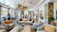 This single-story community clubhouse is defined by a sophisticated blend of natural and contemporary design elements. The entry features large-format tile flooring, a skylight ceiling that floods the space with natural light, floor-to-ceiling glass panels, and prominent stacked-stone columns that establish a warm, resort-inspired aesthetic carried throughout the facility.

The expansive lounge area serves as the centerpiece, showcasing a coffered ceiling with gold-accent cove lighting and striking spiral pendant fixtures. Multiple seating arrangements—including neutral sofas, leather armchairs, and accent chairs—are organized around a linear fireplace and wall-mounted television. Built-in display niches and curated artwork contribute to a polished, gallery-like atmosphere.

Adjacent to the lounge, a well-equipped kitchen provides warm wood cabinetry, light countertops, tile backsplash, and a full suite of appliances beneath a window with outdoor views. A business nook tucked along a corridor offers a dedicated workstation set within a bold blue alcove. The fitness center occupies a bright, vaulted space lined with expansive windows, housing a comprehensive array of cardio and strength equipment. Throughout, recessed lighting, decorative art, and lush greenery reinforce a cohesive and inviting environment.