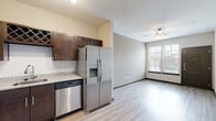 This single-floor residence offers a clean, modern aesthetic highlighted by light hardwood-style flooring that flows seamlessly throughout the entire space. The kitchen is a standout feature, showcasing rich dark-stained cabinetry with sleek hardware, granite countertops, a classic white subway tile backsplash, and a full suite of stainless steel appliances including a side-by-side refrigerator with ice and water dispenser, a dishwasher, an electric range, and an over-the-range microwave. A built-in wine rack adds a refined touch above the cabinetry. The kitchen opens directly into a bright, airy living area illuminated by natural light from large windows fitted with horizontal blinds. A ceiling fan provides year-round comfort in the living space, which also features a charcoal-toned front entry door for a modern contrast against the neutral walls. The bathroom maintains the same design language as the rest of the unit, featuring a dark wood vanity with a granite countertop, a large framed mirror, and a combined shower and soaking tub. A dedicated laundry area with washer and dryer hookups adds everyday convenience. The overall ambiance is fresh, neutral, and move-in ready.