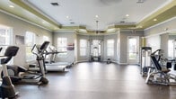 This fitness center offers a spacious, well-appointed environment designed to support a comprehensive workout experience. The room features a distinctive tray ceiling accented with a bold olive-green band and crisp white crown molding, giving the space a polished, upscale character. Recessed lighting and ceiling fans are integrated throughout, ensuring consistent illumination and airflow across the entire area.

The rubber-floored layout is organized to accommodate a wide range of exercise modalities. Cardio equipment, including multiple treadmills, an elliptical, and a stationary bike, lines one section of the room, while a dedicated strength training zone houses cable machines, a multi-station weight unit, a dumbbell rack, and a weight bench. Exercise balls in various sizes are neatly stored on a rack, and wall-mounted mirrors expand the visual space and assist with form during workouts.

Multiple windows with white blinds line the walls, flooding the room with natural light and offering views of the surrounding exterior. A mounted flat-screen television and motivational wall decals contribute to the energetic atmosphere. A recycling station near the entrance underscores a commitment to sustainability. The facility is accessible via glass-paneled doors, providing a welcoming and functional entry point.
