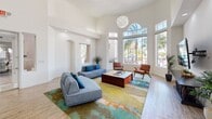 Located in Phoenix, AZ, this single-floor clubhouse offers a well-appointed communal space designed with a modern aesthetic and an inviting atmosphere. The living room is the centerpiece of the space, boasting dramatically soaring ceilings that amplify the sense of openness. Floor-to-ceiling windows flood the room with abundant natural light while framing views of the surrounding palm trees, creating a seamless connection to the Arizona outdoors. A sleek, wall-mounted fireplace with an arched architectural detail above serves as a focal point, flanked by contemporary leather seating. Additional mid-century modern armchairs and a vibrant multicolored area rug add warmth and personality to the lounge area. Light wood-tone flooring runs throughout, complementing the crisp white walls. The kitchen is equally striking, featuring a bold geometric-patterned accent wall in a rich palette of terracotta, teal, gold, and gray. Dark cabinetry with white quartz countertops provides a sophisticated contrast, while a spacious island with bar seating and a Sputnik-style pendant light overhead enhance both functionality and style. Stainless steel appliances complete the well-equipped kitchen. Together, these spaces create a dynamic and welcoming clubhouse environment.