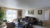 This single-floor residence offers a thoughtfully arranged and welcoming living space. The living room serves as the centerpiece of the home, showcasing a classic and curated interior design aesthetic. A textured popcorn ceiling adds architectural character overhead, while neutral-toned walls provide a warm and understated backdrop throughout. Large windows allow generous amounts of natural light to fill the room, offering views of the surrounding greenery outside.

The living room is furnished with a richly upholstered dark brown sofa set, accented by ornate carved wooden frames that lend an elegant, traditional feel. Decorative throw pillows in complementary earth tones enhance the comfortable seating arrangement. A glass-topped coffee table with a dark wood frame anchors the space atop a bold patterned area rug, adding visual interest to the floor.

The walls are adorned with a curated collection of framed artwork, including a prominently displayed landscape painting in an ornate gold frame, contributing to the room's refined character. Soft, neutral carpeting underfoot adds to the overall sense of warmth and comfort. The cohesive design choices throughout create a space that is both functional and aesthetically inviting, reflecting a classic, well-appointed home interior.