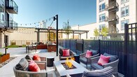 Located in the heart of Durham, NC, this thoughtfully designed outdoor courtyard amenity space offers a dynamic and social environment for residents and guests alike. The courtyard is anchored by a central fire pit lounge area, surrounded by comfortable seating with vibrant accent pillows, making it an ideal gathering spot for relaxed conversation or evening socializing. Overhead string lights create a warm, inviting ambiance throughout the space, complementing the open sky above.

A dedicated outdoor kitchen and grilling station, finished with classic brick detailing, sits alongside outdoor dining tables and chairs, providing a functional space for al fresco meals. Multiple pergola structures define distinct zones within the courtyard, offering partial shade while maintaining an open-air feel. Raised planter beds and carefully curated landscaping add greenery and visual interest throughout.

A lush artificial turf lawn area provides a versatile recreational zone, complete with a cornhole board for casual outdoor games. The surrounding multi-story building facade, featuring a contemporary mix of gray siding, large windows, and private balconies, frames the courtyard and creates an intimate, urban oasis feel. The expansive concrete patio offers ample open space, tying all the amenity zones together seamlessly.