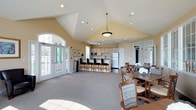This single-floor community clubhouse offers a thoughtfully designed layout that balances social, functional, and recreational spaces. The welcoming entry foyer features tile flooring, a decorative accent table, and transom-topped French doors, with clearly marked access to a reception area and business center.

The grand lounge is a standout architectural feature, boasting soaring vaulted ceilings, expansive arched windows that flood the space with natural light, and a dramatic floor-to-ceiling stacked stone fireplace flanked by leather armchairs. French doors open directly to the outdoor patio, creating a seamless indoor-outdoor connection.

The full kitchen is equipped with light wood cabinetry, granite-style countertops, stainless steel appliances including a dishwasher and microwave, and hardwood flooring. A large peninsula with bar-height seating adds a social element to the kitchen space.

Adjacent dining areas accommodate multiple seating configurations with wood tables and upholstered chairs, complemented by a wall-mounted flat-screen television and views to the surrounding greenery.

Outside, a curved pergola-covered patio provides a gracious setting for outdoor gatherings, overlooking a lush, open lawn framed by mature trees. The combination of refined interior details and inviting communal amenities makes this clubhouse a versatile and welcoming gathering place.