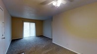 This single-floor residential space offers a straightforward and functional layout designed for comfortable everyday living. The main living area is finished with wall-to-wall carpeting, providing a soft and warm underfoot feel throughout. A ceiling fan with an integrated light fixture is centrally mounted on the textured ceiling, ensuring consistent air circulation and ambient lighting across the room. Neutral-toned walls in a warm beige hue create a calm and versatile backdrop, making the space easy to personalize with a variety of décor styles. A sliding glass door fitted with vertical blinds serves as a prominent architectural feature, allowing natural light to filter into the room while also offering direct access to the outdoors. The door frames a view of the surrounding greenery, adding a refreshing connection to the outside environment. Clean white baseboards line the perimeter of the room, providing a crisp contrast to the carpeted floors. The overall ambiance of the space is understated and move-in ready, with a clean, open feel that lends itself well to flexible furniture arrangements and personal customization.