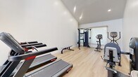 Located at 15123 Brookhurst St in Westminster, CA, this dedicated exercise room offers a clean, modern fitness environment designed to support a comprehensive workout routine. The space features soaring ceilings that create an open, airy atmosphere, complemented by large floor-to-ceiling windows and glass doors that flood the room with natural light and provide views of an outdoor covered pergola and pool area beyond.

The light wood laminate flooring runs throughout, lending warmth and a polished aesthetic to the space. Crisp white walls and recessed track lighting further enhance the bright, energizing ambiance. The room is equipped with a well-rounded selection of fitness equipment, including multiple treadmills, a stationary bike, an elliptical trainer, a multi-function cable and weight machine, and a flat workout bench — offering both cardio and strength training options.

A water fountain is conveniently installed near the entrance, and a posted fitness guide is visible on the wall. Glass doors open directly to the outdoor amenity area, seamlessly connecting indoor exercise with the outdoor recreational space. This thoughtfully designed fitness room provides a focused and inviting setting for residents to maintain an active and healthy lifestyle.