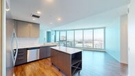 Located along the Long Beach waterfront, this modern 2-bedroom, 2-bathroom residence offers an open, well-appointed layout with stunning panoramic views of the marina, harbor, and surrounding cityscape. Floor-to-ceiling windows flood the living area with natural light, and warm hardwood-style flooring flows throughout the main living spaces. The open-concept kitchen and living room create an inviting, functional hub at the heart of the home. The kitchen is outfitted with sleek gray cabinetry, white quartz countertops, a white tile backsplash, and a full suite of stainless steel appliances including a refrigerator, range, microwave, and dishwasher. A movable kitchen island with built-in wine storage adds both prep space and versatility. The spacious primary bedroom features corner floor-to-ceiling windows framing breathtaking harbor views, plush carpeting, and direct access to a well-appointed en-suite bathroom. The primary bath includes a glass-enclosed shower, a floating vanity with an illuminated mirror, and hardwood-style flooring, with a walk-in closet conveniently adjacent. The second bathroom offers a soaking tub and shower combination with elegant tile surround. A generous walk-in closet with wire shelving provides ample storage throughout.