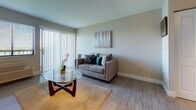 This 2-bedroom, 1-bathroom apartment is located on the fourth floor of Grand Court Apts in Miami, Florida, offering a well-designed layout across a single level. The living area serves as the heart of the unit, featuring wood-style flooring, neutral-toned walls, and generous natural light streaming through a large sliding glass door with vertical blinds that open to an outdoor view. A round glass coffee table and a comfortable loveseat create an inviting, functional living space. Both bedrooms are thoughtfully appointed, each featuring wood-style flooring, individual climate control units, and windows that frame picturesque water views of the surrounding Miami landscape. The bedrooms provide ample space for furniture and storage, with dark wood sleigh bed frames and matching dressers lending a cohesive, classic aesthetic throughout. The overall ambiance of the apartment is calm and bright, with soft neutral wall tones complementing the natural light that flows through every room. The fourth-floor elevation enhances the sense of openness and provides residents with elevated water and skyline views, making this a distinctive and comfortable Miami residence.