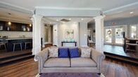 Located at 2222 Ashley River Rd in Charleston, SC, this single-floor space showcases a thoughtfully designed interior that blends classic Southern charm with modern sophistication. Rich hardwood flooring flows throughout, tying together a cohesive and inviting aesthetic.

The kitchen is a standout feature, appointed with dark espresso cabinetry — including glass-front upper cabinets — set against a shimmering mosaic tile backsplash. Light quartz countertops provide striking contrast, while stainless steel appliances and pendant lighting add a contemporary edge. A generous island offers additional prep and gathering space, making this kitchen as functional as it is stylish.

The open living area is anchored by decorative white columns that define the space without closing it off. A bold navy accent wall frames a tiled fireplace and mounted television, creating a dramatic yet elegant focal point. Multiple windows allow abundant natural light to pour in, offering views of the surrounding Charleston landscape. Crown molding throughout lends an air of refinement, complementing the recessed lighting overhead.

The overall ambiance is one of polished comfort — a space that balances architectural detail with warm, livable design, ideal for both relaxed everyday use and sophisticated entertaining.