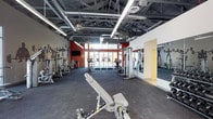 This fitness center offers a comprehensive workout environment designed to accommodate a wide range of exercise needs. The space features an open, industrial-style layout with exposed ductwork and steel trusses overhead, lending the facility a modern, energetic atmosphere. Large windows along the far wall allow generous natural light to fill the floor, creating a bright and motivating environment.

The facility is well-equipped with a robust selection of both cardio and strength training machines. Treadmills and stationary bikes are arranged along the perimeter, offering excellent options for cardiovascular workouts. A variety of cable machines, weight benches, and multi-station strength equipment are thoughtfully distributed throughout the space, allowing multiple users to train simultaneously without congestion.

Dark rubber flooring covers the workout area, providing both durability and safety underfoot. A flat-screen television is mounted on a bold red accent wall, adding an entertainment element for members during their sessions. Floor-to-ceiling mirrors on one wall enhance the sense of space while allowing users to monitor their form. The combination of industrial design elements, practical amenities, and varied equipment makes this fitness center a functional and well-appointed space for users of all fitness levels.