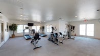This residential community offers a comprehensive range of amenities designed to support an active and social lifestyle. The indoor fitness center is bright and well-lit, featuring recessed lighting, a textured ceiling, and large windows that invite natural light. It is equipped with cardio machines including treadmills, an elliptical, and a stationary bike, as well as a multi-station strength training unit — providing a thorough workout experience without leaving the property.

Outdoors, the centerpiece is a generously sized swimming pool with crystal-clear turquoise water, framed by tall swaying palm trees that lend a resort-like ambiance. A poolside patio features lounge chairs for relaxation, along with a covered pergola supported by stone columns, offering shaded seating with bistro-style furniture and a vibrant red umbrella.

The community's recreational spaces extend further with a full outdoor basketball court, complete with dual hoops and bench seating. A dedicated children's play area includes a colorful multi-level playground structure with slides and activity panels, as well as a separate swing set — both set on wood chip surfacing for safety. Picnic tables with red umbrellas provide casual outdoor dining spots. The surrounding buildings feature a distinctive Southwest-inspired architecture with warm yellow and earth-tone exteriors and red tile roofing.