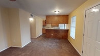 This single-floor unit offers an open-concept layout that seamlessly connects the kitchen and living area, creating a functional and cohesive living space. The kitchen is equipped with warm-toned wooden cabinetry extending to the upper wall, providing ample storage. A built-in peninsula separates the kitchen from the living area, offering additional counter space and serving as a natural divider between the two zones. A white refrigerator and a sink integrated into the counter complete the kitchen's essential features.

The living area is finished with wall-to-wall carpeting in a neutral tan tone, contributing to a comfortable and understated atmosphere. The walls are painted in a light cream and yellow palette, adding warmth to the interior. Natural light enters through a window fitted with blinds on the adjacent wall, brightening the space during the day. A flush-mount ceiling light fixture provides overhead illumination. A doorway visible toward the rear of the space leads to an additional room or hallway, suggesting further functional areas within the unit. An overhead HVAC vent is also present, indicating a central air system.
