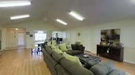 This single-floor space offers an open-concept layout that seamlessly connects the living and dining areas, creating a welcoming and functional environment. The living area is anchored by a generously sized sectional sofa in warm olive tones, accented with decorative green and patterned throw pillows, and centered around a dark coffee table. The layout provides ample seating and encourages comfortable gatherings. Vaulted ceilings rise overhead, enhancing the sense of openness, while a ceiling fan ensures year-round comfort. Bright fluorescent overhead lighting keeps the space well-illuminated. The dining area is situated near a window that allows natural light to filter in, featuring a dining table with chairs ideal for group meals. Hardwood-style flooring flows throughout, adding warmth and cohesion to the space. A dark wood media console or sideboard is positioned along one wall, offering additional storage and display space. Several doorways lead to adjoining rooms, suggesting additional functional spaces beyond the main open area. The overall ambiance is casual, practical, and inviting, making it well-suited for communal or shared use.