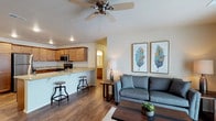 This single-floor space showcases an open-concept layout that seamlessly connects the living area and kitchen, creating a cohesive and functional environment. The living area features rich, dark hardwood-style flooring that extends throughout, anchoring the space with warmth and continuity. A ceiling fan is centrally positioned overhead, providing both practical airflow and a subtle design element.

The kitchen is equipped with warm honey-toned cabinetry, stainless steel appliances including a microwave and range, and light granite-style countertops that add a touch of elegance. A raised breakfast bar with industrial-style bar stools serves as both a casual dining option and a natural dividing element between the kitchen and living spaces.

The living area is bright and welcoming, with neutral-toned walls that complement the contemporary furnishings. Recessed lighting and a floor lamp contribute to a well-lit ambiance throughout. Decorative botanical artwork adds a refreshing accent to the walls. A hallway visible from the main area suggests additional rooms beyond, hinting at further functional spaces within this well-designed, single-level layout.