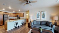 This single-floor space showcases an open-concept layout that seamlessly connects the living area and kitchen, creating a cohesive and functional environment. The living area features rich, dark hardwood-style flooring that extends throughout, anchoring the space with warmth and continuity. A ceiling fan is centrally positioned overhead, providing both practical airflow and a subtle design element.

The kitchen is equipped with warm honey-toned cabinetry, stainless steel appliances including a microwave and range, and light granite-style countertops that add a touch of elegance. A raised breakfast bar with industrial-style bar stools serves as both a casual dining option and a natural dividing element between the kitchen and living spaces.

The living area is bright and welcoming, with neutral-toned walls that complement the contemporary furnishings. Recessed lighting and a floor lamp contribute to a well-lit ambiance throughout. Decorative botanical artwork adds a refreshing accent to the walls. A hallway visible from the main area suggests additional rooms beyond, hinting at further functional spaces within this well-designed, single-level layout.