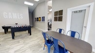 This single-floor amenity space is thoughtfully designed to accommodate multiple activities within a cohesive, open layout. The main area features light wood-look flooring throughout, vaulted ceilings with recessed skylight panels that flood the space with bright, even lighting, and neutral gray walls that create a clean, contemporary backdrop.

The recreational zone includes a foosball table, offering a fun focal point for social gatherings. Adjacent to it, a dining area is furnished with a dark rectangular table and bold blue metal chairs, providing a functional space for meetings or casual dining. A display shelf along one wall showcases a series of framed artwork, adding a curated, decorative touch to the space.

A open passageway connects the main area to a dedicated fitness room, which is equipped with cardio machines including ellipticals and stationary bikes, visible through the opening. A grid-pattern wooden wall accent decorates the shared wall between the two zones, adding texture and visual interest.

Overall, the space balances practicality and comfort, making it well-suited as a community or shared amenity facility.
