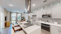 This well-appointed single-floor residence offers a functional and stylish layout comprising a kitchen, living room, bedroom, and closet. The open-concept kitchen is a standout feature, anchored by a large center island with a built-in gas cooktop, striking stainless steel hood vent, and seating for multiple guests. White shaker-style cabinetry reaches ceiling height with frosted glass upper accents, complemented by a tile backsplash, quartz countertops, and a full suite of stainless steel appliances including a built-in oven, microwave, and French door refrigerator. Light wood-tone flooring flows throughout, unifying the space. The living room is bright and inviting, with large windows that draw in generous natural light and offer outdoor views. A warm wood slat accent wall adds texture and visual interest to both the living area and bedroom. The bedroom is a comfortable retreat, featuring large windows with outdoor views, wood-plank flooring, and a ceiling fan. A dedicated closet provides practical storage. Throughout the residence, a cohesive neutral palette, clean architectural lines, and modern lighting fixtures create a bright, contemporary ambiance that feels open and livable.