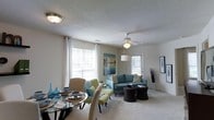 Located at 381 Darlington Avenue in Wilmington, North Carolina, this single-story residence offers a well-designed, open-concept layout that seamlessly connects the living and dining areas. The combined living and dining space is bright and welcoming, benefiting from multiple windows dressed with white curtains that allow generous natural light to fill the room. The living area features plush carpeting underfoot, a ceiling fan with integrated lighting, and neutral-toned walls that create a versatile backdrop for a range of décor styles. A teal sofa anchors the seating area, complemented by accent chairs in green and cream tones, rounded out by wicker-style coffee tables that add a casual, coastal feel consistent with the Wilmington setting. The dining area, positioned adjacent to the living space, includes a glass-top table with cushioned seating, creating a practical yet stylish area for meals. Coastal-themed artwork and decorative accents throughout contribute to the overall warm and inviting ambiance. The textured ceiling and flush-mount lighting add to the finished character of the interior, making this a comfortable and functional single-floor residence.