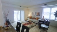 This single-floor space offers a comfortable and well-organized layout designed for everyday living. The living area serves as the centerpiece of the space, featuring plush carpeting underfoot and a warm, neutral color palette that creates a relaxed and inviting atmosphere. A large sliding glass door floods the room with abundant natural light and provides direct access to an outdoor balcony, offering pleasant exterior views. Vertical blinds frame the door, allowing for flexible light control throughout the day. The living room is thoughtfully arranged with a generous sofa accented by earth-toned throw pillows, complemented by accent chairs with studded detailing that add a touch of character. A glass-top coffee table with a sleek metal frame anchors the seating area over a multi-colored area rug, adding visual depth to the space. Decorative framed artwork adorns the walls, contributing to the curated, homey feel of the interior. A secondary window on the adjacent wall provides additional natural light, enhancing the bright and airy quality of the room. The overall ambiance is warm, functional, and welcoming, making it a comfortable retreat suited for both relaxing and entertaining.
