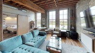 Located at 331 S 3rd St in Milwaukee's Walker's Point neighborhood, this single-floor loft showcases a compelling blend of historic industrial architecture and contemporary design. The space retains striking original features throughout, including exposed cream brick walls, raw concrete surfaces, and overhead timber beam ceilings with visible joists that lend a distinctly urban, warehouse-style character.

The open living area is generously scaled, anchored by tall multi-pane windows that flood the room with natural light and frame views of the surrounding cityscape. Original hardwood floors run throughout, adding warmth against the raw structural elements. The living room provides ample space for comfortable seating and entertainment, with the exposed brick wall serving as a dramatic backdrop for a wall-mounted television.

The kitchen features white cabinetry, stainless steel appliances, open wooden shelving with subway tile backsplash, and a large island with light countertops — a functional yet stylish contrast to the industrial surroundings.

The bedroom areas maintain the loft's aesthetic with exposed brick, timber ceilings, and large windows offering outdoor views. Built-in shelving provides practical storage while contributing to the overall design.

The bathroom offers a clean, modern contrast with light wood-toned cabinetry, white countertops, a walk-in glass shower, and neutral tile flooring, delivering a serene, contemporary finish.