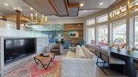 This well-appointed clubroom is a single-floor community gathering space that seamlessly blends rustic and contemporary design elements throughout its multiple distinct zones. The main lounge area anchors the space with a plush sofa, cushioned armchairs, and a bold patterned area rug, all centered around a reclaimed wood accent wall displaying vibrant artwork. Coffered ceiling details with warm wood inlays and a striking gold chandelier add architectural interest overhead, while expansive windows flood the area with natural light and frame views of the outdoor pool and palm trees beyond. An elegant dining area adjoins the lounge, furnished with an oval dark-topped table and upholstered chairs beneath a matching chandelier. A dedicated café station features white cabinetry, a coffee bar, wall-mounted television, pendant lighting, and a communal wooden high-top table set against a textured honeycomb tile backsplash. A separate lounge nook offers built-in bench seating with colorful mosaic tile accents, paired with round pedestal tables and cylindrical stools under decorative wall sconces. A dedicated game room, accessed through glass-paneled French doors, houses a billiards table and additional recreational space. Throughout, geometric patterned flooring, curated artwork, and eclectic furnishings create a vibrant, inviting atmosphere designed for community engagement and relaxation.