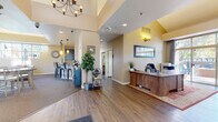 This well-appointed single-floor community space is designed to be both functional and inviting, offering a variety of areas that cater to residents and visitors alike. The entry reception area sets the tone immediately, featuring a warm, rustic wood front desk accented by wall sconces and a framed window view, all beneath soaring vaulted ceilings that enhance the sense of openness. Rich hardwood flooring flows throughout the main areas, anchored by a striking wrought-iron chandelier overhead.

Adjacent to the reception, a fully equipped kitchenette provides dark espresso cabinetry, granite countertops with a mosaic tile backsplash, stainless steel appliances including a microwave and compact refrigerator, and pendant lighting — all opening toward the communal lounge and dining area with bar-height seating.

The lounge is a standout feature, bathed in natural light from an expansive wraparound window arrangement that frames outdoor views. A corner stacked-stone fireplace with a mounted television serves as a focal point, complemented by comfortable leather and upholstered seating arranged around a rustic wood coffee table.

A private office or business nook offers a workstation with seating, warm carpeting, a natural wood built-in storage unit, and a window with outdoor views. The space is completed by an accessible restroom finished with tile walls, wood-look flooring, and ADA-compliant fixtures.