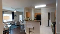 This single-story residence showcases an open-concept layout that seamlessly connects the kitchen and living areas, creating a cohesive and functional living environment. The kitchen is equipped with warm honey-toned cabinetry, dark granite-style countertops, and black appliances, including a full-size refrigerator. A peninsula with an overhanging breakfast bar provides additional seating with sleek white bar stools, making it an ideal space for casual dining or entertaining. Recessed and overhead lighting ensures the kitchen remains well-lit throughout the day.

The flooring transitions from dark hardwood in the living area to lighter tile in the kitchen, clearly delineating the two spaces while maintaining a cohesive aesthetic. Crown molding runs throughout the space, adding a refined architectural detail to the standard-height ceilings. A ceiling fan in the living area enhances comfort and air circulation. The living area is carpeted with a teal area rug and furnished with neutral-toned seating, while a window near the entryway allows natural light to filter in, brightening the overall ambiance of the space.