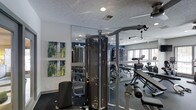 This fitness center offers a comprehensive workout environment designed to accommodate a variety of exercise routines. At its centerpiece stands a large multi-station cable machine, providing users with versatile strength-training options. The space is thoughtfully arranged to maximize functionality, with additional equipment including a weight bench with plates and various resistance machines visible throughout the room.

Full-length mirrors line one wall, visually expanding the space while allowing users to monitor their form during workouts. The flooring features dark, durable carpeting suited for a high-traffic fitness environment. Recessed lighting in the textured white ceiling provides even illumination throughout the room, complementing the natural light that filters in through a window visible in the mirrored reflection.

The walls are painted a deep navy blue on select surfaces, creating a bold contrast against the lighter walls adorned with a set of abstract green and yellow canvas artworks, adding a touch of color and visual interest to the athletic setting. The overall ambiance is practical yet polished, offering a well-maintained facility suitable for both casual and dedicated fitness enthusiasts.