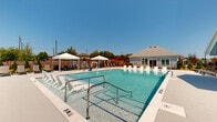 Located at 500 NW E Crawford Dr in Fate, TX, this single-story community amenity facility spans a generously sized footprint designed for outdoor recreation and relaxation. At its centerpiece is a large, sparkling resort-style swimming pool with clear blue water, accessible via stainless steel handrails and surrounded by an expansive pool deck. The deck is well-appointed with rows of white lounge chairs and shaded canopy umbrellas, providing ample space for sunbathing and leisure. A dedicated shallow end is clearly marked, making the pool suitable for a range of swimmers.

Adjacent to the pool, a covered pavilion with a clean white metal roof shelters an outdoor seating area furnished with plush lounge furniture, ideal for gathering and entertaining. A built-in grill station nearby adds convenience for outdoor cooking. The grounds extend into a neatly manicured lawn area dotted with mature shade trees and Adirondack-style chairs, offering a tranquil retreat. A dedicated game lawn features cornhole boards for casual recreation.

Thoughtfully landscaped pathways with ornamental grasses, mulched beds, and native plantings connect the various zones throughout the property. Black iron fencing defines the perimeter while maintaining an open, airy feel. The entire facility is bathed in natural Texas sunlight, creating a vibrant, welcoming outdoor environment.