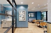 Located at 2031 Westcreek Ln in Houston, TX 77027, this thoughtfully designed 580-square-foot single-floor amenity space blends modern aesthetics with functional versatility across two distinct areas. The main lounge and dining area showcases light oak herringbone flooring, tray ceilings with recessed lighting, and an eye-catching terrazzo-patterned accent wall. Warm golden velvet chairs surround sleek dark marble-topped tables, complemented by a teal feature wall adorned with oval mirrors and a cushioned bench. A built-in credenza with light wood cabinetry provides coffee and beverage service. A large wall-mounted display screen and additional built-in cabinetry enhance the space's functionality. The second area features a glass-enclosed conference room with floor-to-ceiling black steel-framed partitions, offering full visibility while maintaining acoustic separation. Inside, a round conference table with ergonomic chairs sits atop dark marble-style flooring. Lush potted tropical plants add natural warmth throughout both areas, while abundant natural light filters in through exterior-facing glass walls, creating a bright, professional, and inviting atmosphere.