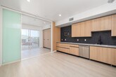 Located at 1140 S Normandie Ave in Los Angeles, this modern one-bedroom, one-bathroom unit is designed with a clean, contemporary aesthetic throughout. Light wood-tone finishes and pale luxury vinyl plank flooring run consistently across the space, creating a cohesive and bright atmosphere. A long hallway with recessed lighting and built-in wood cabinetry leads from the entry into the main living area. The open-plan kitchen and living room is a functional centerpiece, featuring flat-front light wood cabinetry, striking dark mosaic tile backsplash, white quartz countertops, and a full suite of stainless steel appliances including a range, microwave, dishwasher, and refrigerator. Sliding glass doors open directly from the living area onto a private balcony enclosed by sleek metal railings, offering open views of the surrounding Los Angeles neighborhood under sunny skies. The bedroom is a well-proportioned retreat served by two closets for ample storage. The bathroom is finished in the same light, modern palette with a soaking tub, wood-framed vanity with white countertop, a large mirror with black trim, and a built-in open shelving tower. An in-unit laundry area adds everyday convenience to this thoughtfully designed urban residence.