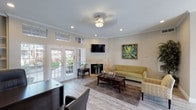 Located at 8403 Williamcrest Ln in Houston, TX, this single-story space presents a well-organized and inviting layout designed for both comfort and functionality. The interior is finished with light wood-tone flooring throughout, crisp crown molding, and neutral walls that create a warm, professional atmosphere.

The main lounge area serves as the heart of the space, anchored by a tile-surround fireplace with a wall-mounted flat-screen television above it. A tufted olive-green sofa, patterned accent chair, and a dark wood coffee table are arranged over a decorative area rug, lending a polished yet approachable feel. Recessed lighting and a ceiling fan with a light fixture ensure the area is well-lit and comfortable. A large framed botanical artwork adds a notable decorative touch to the room.

French doors along multiple walls invite abundant natural light and provide views of the exterior. Transom windows positioned above the doors further enhance the brightness of the interior.

A dedicated office area features a substantial dark wood desk, built-in shelving, and seating for guests, making it well-suited for meetings or administrative use. The space flows openly from the lounge into this functional zone, reinforcing an overall sense of openness and cohesion throughout the layout.